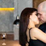 A man and a woman embrace and kissing. Bald guy in a suit and a girl in a black dress in a restaurant, loft. Valentine’s Day. Banner